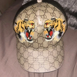 Gucci keps med tigertryck och mesh - Snygg Gucci keps med klassiskt GG-mönster i beige och brunt, prydd med två stora broderade tigrar framtill. Kepsen har svart mesh på sidorna och justerbar rem bak med röd-gröna detaljer. Perfekt statement-piece för dig som vill sticka ut. Och box och påse ingår med beställning. Pris kan diskuteras 