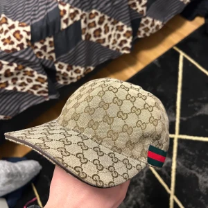 Beige keps från Gucci - Snygg beige keps från Gucci med det ikoniska GG-mönstret. Kepsen har en justerbar rem baktill och en grön och röd randdetalj på sidan. Perfekt för att ge din outfit en lyxig touch.