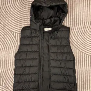 Snygg svart dunväst från H&M med huva och dragkedja. Perfekt för lager-på-lager under kyliga dagar. 