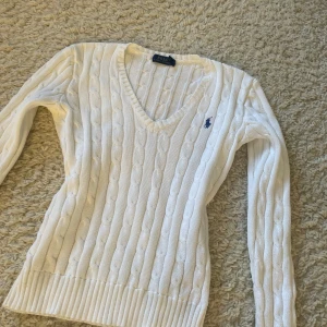 Vit stickad tröja från Polo Ralph Lauren - Säljer en stilren vit stickad tröja från Polo Ralph Lauren med v-ringning och det klassiska logotypbroderiet på bröstet. Tröjan har en ribbad struktur och långa ärmar, perfekt för en elegant och avslappnad look.