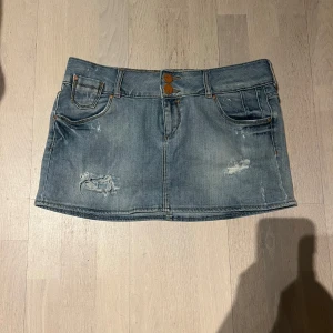 Jeanskjol från Bershka - Snygg jeanskjol från Bershka i en klassisk blå denimfärg. Kjolen har en kort längd med slitna detaljer och två knappar framtill. Den har också praktiska fickor både fram och bak. Perfekt för en avslappnad stil. Lågmidjad 