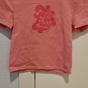Rosa t-shirt från Bik Bok - Säljer en rosa t-shirt från Bik Bok med trycket 'Take it Easy' i en cool, retro stil på framsidan. T-shirten är kortärmad och gjord i mjukt bomullsmaterial. Perfekt för en avslappnad look!