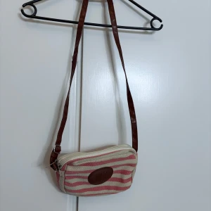 Randig axelväska med brunt läderband - Säljer en söt axelväska med rosa och beige ränder. Väskan har ett brunt läderband och en dragkedja upptill. Perfekt för en avslappnad dag ute.