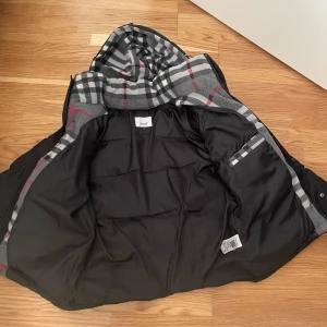 Svart pufferjacka från Burberry - Säljer en snygg svart pufferjacka från Burberry med klassiskt rutigt foder. Jackan har en huva och stängs med dragkedja och knappar. Perfekt för kyliga dagar! Den passar s och m, jätteskön och fett fin. Kan sänka vid en snabb äffar för 1300