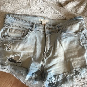 Ljusa jeansshorts från H&M - Säljer ett par ljusa jeansshorts från H&M med slitna detaljer och fransar vid benen. Perfekta för sommardagar. Midjemått: ca 38cm Shortsen har en klassisk femficksdesign och knappgylf.
