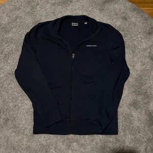 Fullzip Jack & Jones - Säljer en stilren mörkblå zip från Jack & Jones med dragkedja framtill. Perfekt för en avslappnad look. Den har långa ärmar och en diskret logga på bröstet. Passar bra till både jeans och joggers.