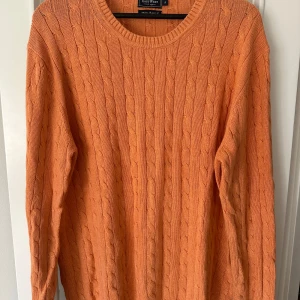 Orange kabelstickad tröja från East West - Säljer en snygg orange kabelstickad tröja från East West. Tröjan har ett klassiskt kabelstickat mönster och rund halsringning. 