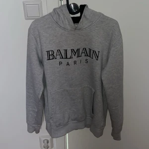 Grå hoodie från Balmain - Säljer en stilren grå hoodie från Balmain Paris. Tröjan har en klassisk design med en stor ficka framtill och Balmain-loggan tryckt i svart på bröstet. Perfekt för en avslappnad och trendig look.