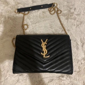 Svart quiltad axelväska från YSL - Snygg svart quiltad axelväska med guldfärgade kedjedetaljer och logotyp. Väskan har en elegant design med en justerbar axelrem och är perfekt för att lyfta vilken outfit som helst.