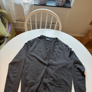 Grå kofta från Polo Ralph Lauren - Säljer en stilren grå kofta från Polo Ralph Lauren. Koftan har en v-ringad design med knappar framtill och långa ärmar. Perfekt för en klassisk och avslappnad look.