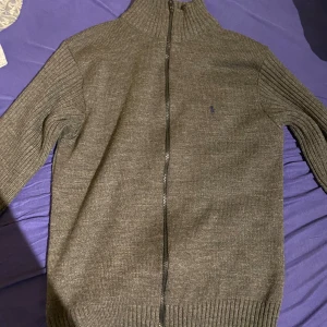 Grå stickad tröja från Ralph Lauren - Säljer en stilren grå stickad tröja från Ralph Lauren med dragkedja framtill. Tröjan har långa ärmar och en ribbad krage.