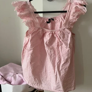 Rosa blus från Vero Moda - Söt rosa blus från Vero Moda med volangdetaljer på axlarna. Blusen har en luftig passform och är perfekt för varmare dagar. Den är tillverkad i ett lätt och bekvämt material.