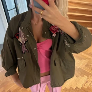 Olivgrön jacka med broderade detaljer - Snygg olivgrön jacka med broderade fåglar och blommor. Jackan har en avslappnad passform med dragkedja och knappar framtill. Perfekt för en cool och avslappnad stil. OBS jackan har två fläckar på baksidan är inte säker om de går bort men man kan testa 🤗tycker dock inte att man lägger så mycket märke till dem 