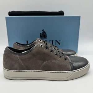 Grå sneakers från Lanvin - Snygga grå sneakers från Lanvin i mocka med snörning och en kontrasterande mörkgrå tå. Skorna har en vit sula som ger en stilren look. Perfekta för en avslappnad men elegant stil.