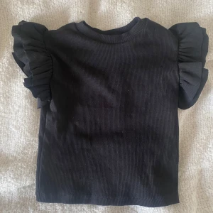 Svart ribbad topp från Gina Tricot.  - En super fin svart ribbad topp från Gina Tricot med volangdetaljer på ärmarna. Perfekt för en vår eller sommar dag. Inga fläckar. Inga hål. Aldrig använd men lappen är tyvärr borta. Storlek 134/140.