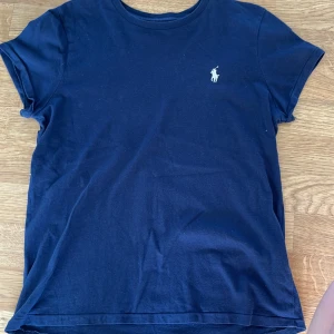 Mörkblå t-shirt från Ralph Lauren - Säljer en klassisk mörkblå t-shirt från Ralph Lauren med det ikoniska broderade logotypen på bröstet. T-shirten är kortärmad och tillverkad i mjuk bomull för en bekväm passform. Perfekt för en stilren och avslappnad look.