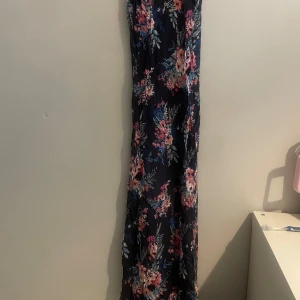 Blommig långklänning med smala axelband - Snygg långklänning med blommigt mönster i rosa, blått och vitt på en mörk bas. Klänningen har smala axelband och en v-ringad design som ger en elegant look.