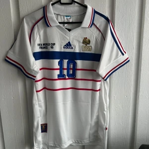 Vit fotbollströja från Adidas - Säljer en vit fotbollströja från Adidas med blå och röda detaljer. Tröjan har korta ärmar och nummer 10 på framsidan samt namnet Zidane på baksidan. Perfekt för fotbollsfans!