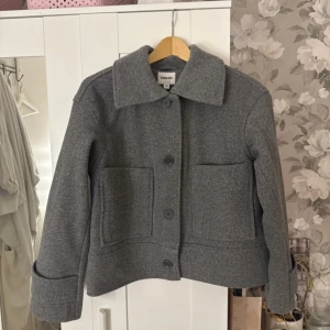 Grå kappa från Kappahl - Säljer en stilren grå kappa från Kappahl i storlek XS. Jackan har en klassisk krage och stora fickor framtill. Perfekt för kyligare dagar med sin tidlösa design och bekväma passform. Använd 1en gång. Original pris 900kr