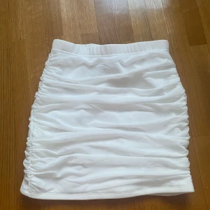 Vit tight kjol med rynkade detaljer - Snygg vit tight kjol med rynkade detaljer på sidorna. Perfekt för en stilren look. Kjolen är kort och har en elastisk midja för bekväm passform.