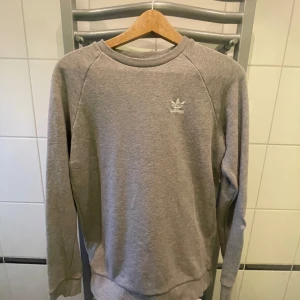 Grå tröja från Adidas - Säljer en stilren grå tröja från Adidas med deras ikoniska logga på bröstet. Tröjan har långa ärmar och en rund halsringning, perfekt för en avslappnad look. Tillverkad i ett mjukt material som ger en bekväm passform.