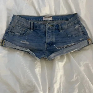 Blå jeansshorts från One Teaspoon - Säljer ett par snygga blå jeansshorts från One Teaspoon. De har en avslappnad passform med hög midja och slitna detaljer för en cool look. Shortsen har en knappgylf och uppvikta benslut. Perfekta nu till sommaren❣️kom privat om du har några frågor, pris går att diskutera 
