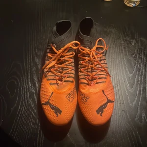 Orange och svarta knappt andvända Puma skor - Ett par puma future fuzionfit evoknit pro acz elite orange. Snygga fotbollsskor från Puma i en iögonfallande orange och svart färgkombination. Skorna har snörning och en hög ankelkrage för extra stöd. Perfekta för fotbollsplanen med sina greppvänliga dobbar.