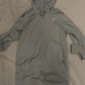 Grå hoodie från Gymshark - Säljer en stilren grå hoodie från Gymshark. Tröjan har en justerbar huva med snörning och en diskret logga på bröstet. Perfekt för både träning och avslappnade dagar. Inga defekter och är som ny
