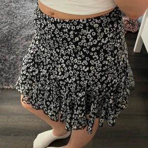 Svart blommig kjol - Snygg svart kjol med vita blommor och volangdetaljer. Kjolen har en bekväm passform💗