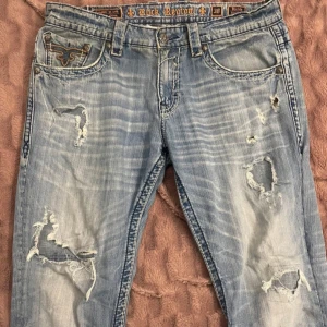 Blå jeans från Rock Revival - Säljer ett par blå jeans från Rock Revival som jag köpte för ca 1 månad sedan. Jag säljer dem då dem var för stora för min smak. Jeansern har slitna detaljer och broderade bakfickor. Dem har en bootcut/loose fit passform. Om priset inte passar kom med ett förslag. (Inga skambud helst)