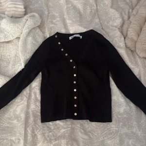 Svart ribbad tröja från Zara - Svart ribbad tröja från Zara med snygg v-ringning och guldiga knappar längs hela framsidan. Långärmad och i mjukt, stretchigt material som sitter skönt på kroppen. Perfekt att styla med jeans eller kjol för en clean look.
