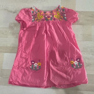 Rosa kortklänning med broderi - Supersöt rosa kortklänning med broderade blommor och fjärilar vid halsringningen och på fickorna. 