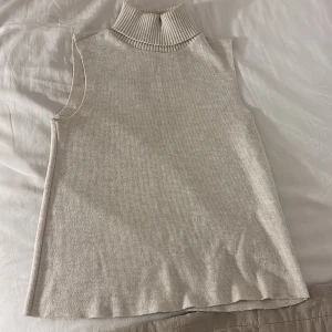 Beige stickad polotopp ärmlös - Säljer en  beige stickad polotopp med hög ribbad krage. Toppen är ärmlös och har en rak passform. Perfekt att styla lager på lager eller bära som den är för en clean look.