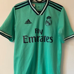 Real Madrid turkos fotbollströja Adidas - Snygg Real Madrid fotbollströja från Adidas i turkos med mörkblå detaljer och klubbmärke på bröstet. Tröjan har korta ärmar, V-ringad krage med ränder och är tillverkad i lätt, andningsbart Climalite-material. Perfekt för dig som älskar fotboll och vill sticka ut på planen.
