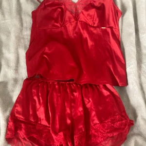 Röd satin linnetopp med spetsdetalj - Snygg röd linnetopp i satin med tunna axelband lopp och v-ringning dekorerad medbv spets. Toppens glansiga material ger en lyxig känsla och den är perfekt för dig som gillar en feminin och trendig stil.