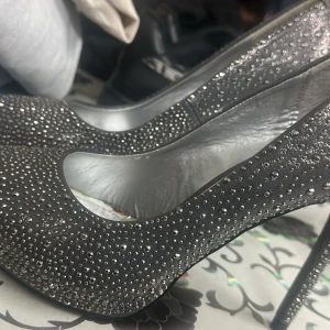 Klackar - Snygga svarta pumps med hög klack och täckt av små glittriga stenar för extra bling. Skorna har rund tå och insida i metallic-silver. Perfekta för dig som vill sticka ut med en glamorös look.