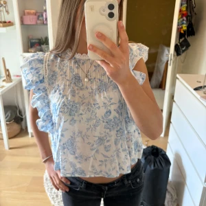 Blommig volangblus från Gina Tricot - Supersöt blus från Gina Tricot i vitt med blått blommönster. Toppen har volangdetaljer längs axlarna, spetskant vid halsen och är ärmlös. Perfekt för en fräsch och romantisk look under varmare dagar.