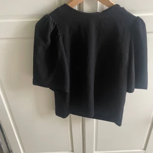 Svart blus med puffärm från H&M - Svart blus från H&M i storlek 38 med korta puffärmar och diskret struktur i tyget. Blusen har rund halsringning och dragkedja bak för enkel påklädning. Perfekt för dig som gillar stilrena och trendiga plagg med lite extra volym i ärmarna.
