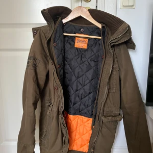 Olivgrön parkajacka från Superdry - Säljer en olivgrön parkajacka från Superdry med broderad logga på ärmen, flera fickor framtill och quiltat foder i svart och orange. Jackan har både dragkedja och knappar, samt en hög krage och huva. Perfekt för kalla dagar och riktigt snygg streetstil. Hittar inte vart storleken står men skulle säga att det är M-L