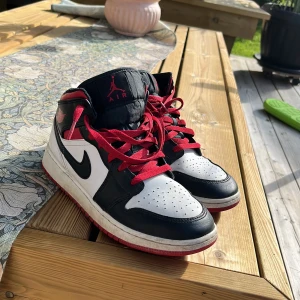 Nike Air Jordan 1 Mid svart/vit/röd - Säljer ett par Nike Air Jordan 1 Mid i klassisk svart, vit och röd färg. Skorna har röda snören, svart swoosh och röd yttersula. Tillverkade i skinn med perforerad tåbox och ikoniska Air Jordan-detaljer på sidan och plös.