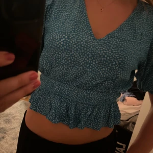 Blommig blå croppad blus från Bikbok - Supersöt blå blus från Bikbok med små vita blommor, v-ringning och korta puffärmar. Modellen är croppad med volangkant nedtill och smock i midjan. Perfekt för dig som gillar en feminin och trendig stil.