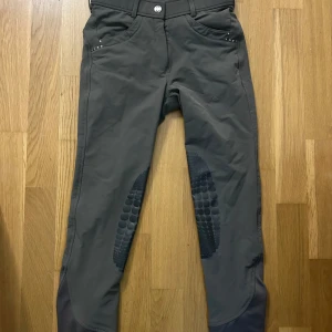 Mörkgrå ridbyxor från CRW med detaljer - Snygga mörkgrå ridbyxor från CRW med silikongrepp på knä och säte, dragkedjefickor bak och små nitar som detalj. Byxorna har hög midja och är tillverkade i ett stretchigt syntetmaterial för bästa komfort i stallet. Lite noppriga i rumpan men inget som syns vid användningen 💘Pris kan diskuteras. 