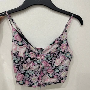 Blommig croppad linnetopp med snörning - Superfin croppad linnetopp från H&M med blommigt mönster i rosa, lila och vitt på svart botten. Toppen har smala axelband, v-ringning och snygg snörning framtill. Perfekt för sommarens alla tillfällen och ger en riktigt söt vibe.