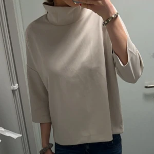 Beige polotröja med trekvartsärm - Säljer en stilren beige polotröja med lös passform och trekvartsärm. Tröjan har hög krage och är tillverkad i ett mjukt material som känns skönt mot huden. Perfekt för dig som gillar minimalistisk och clean stil.