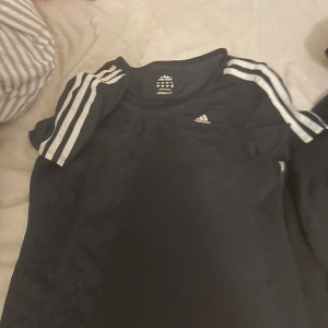 Svart Adidas t-shirt med vita ränder - Svart t-shirt från Adidas med klassiska vita ränder längs axlarna och det ikoniska loggan på bröstet. Kortärmad modell i lätt och sportigt material, perfekt för träning eller chill dagar. Snygg och enkel att matcha med det mesta.