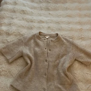 Stickad kofta  - Supermysig beige stickad kofta från Gina Tricot med korta ärmar och rund hals. Koftan har ribbad struktur och knäpps med knappar framtill. Perfekt att slänga över en topp för en chill och stilren look.