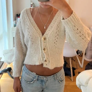 Beige croppad kofta med knappar - Superfin beige croppad kofta med stora träknappar och v-ringning. Stickad i ett mjukt material med snygga håldetaljer på ärmarna och framstycket. Perfekt till jeans för en chill och trendig look.