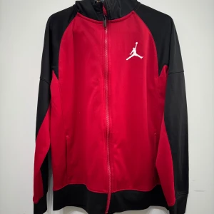 Röd och svart Jordan vindjacka - Snygg röd och svart  kofta från Jordan med hel dragkedja och vit logga på bröstet. Jackan har lång ärm, hög krage och sportig look. Perfekt för dig som gillar streetwear och vill sticka ut med klassisk Jordan-stil.