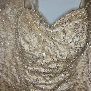 Champagnefärgad långklänning med pärlor - Magisk långklänning i champagnefärgad satin med glittriga paljetter och pärlor över hela klänningen. Hjärtformad urringning och transparenta långa ärmar ger en lyxig känsla. Perfekt för bal eller fest där du vill glänsa extra mycket.