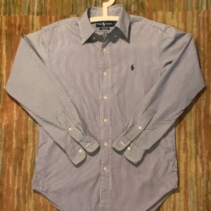 Randig skjorta från Ralph Lauren S - Klassisk blåvit randig skjorta från Ralph Lauren i storlek S. Skjortan har lång ärm, knappar framtill och broderad logga på bröstet. Classic fit-modell i mjuk bomull, perfekt till jeans eller chinos. Snygg krage och stilren design.